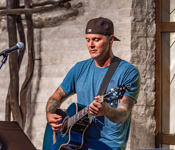 Musician in a blue t-shirt and backward brown cap playing a blue acoustic guitar on a stage with a microphone nearby and a stone wall backdrop.
